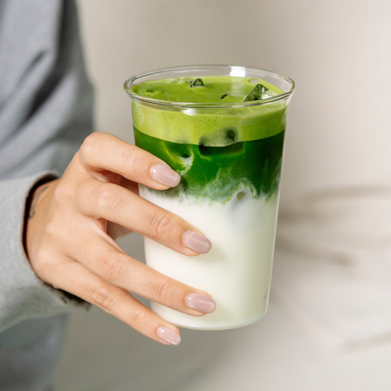 Hand holding a glass of layered green and white drink against a neutral background